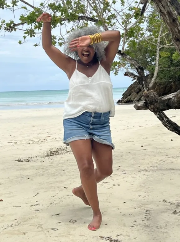 Tami dancing barefoot on a beach, laughing with her arm across her face